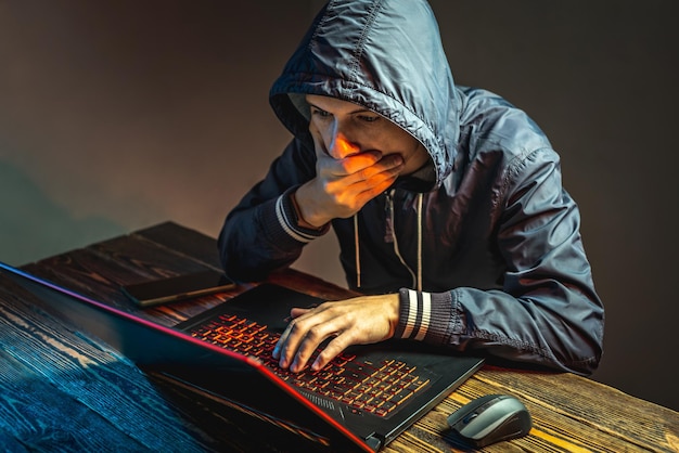 전화가 있는 후드에 있는 해커가 사이버 전쟁 및 Dos 공격의 개념인 어두운 방에서 노트북 키보드에 입력하고 있습니다