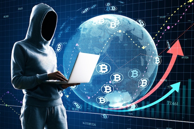 Hacker in hoodie using laptop with glowing globe hologram with financial arrows and money signs on blurry background digital world hacking cryptocurrency banking trade and technology concept