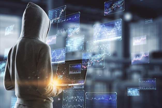 Hacker in hoodie using laptop with various forex screens on blurry office interior background Cryptocurrency hacking bticoin trading and finance concept Double exposure