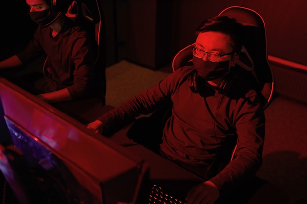 Hacker in beschermend masker zittend op een stoel voor computermonitor werken met nieuwe software in donkere kantoor