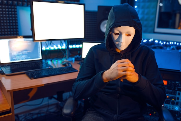 Hacker in mask and hood sitting at his workplace with laptops and PC, information hacking. Internet spy, illegal lifestyle, risk job