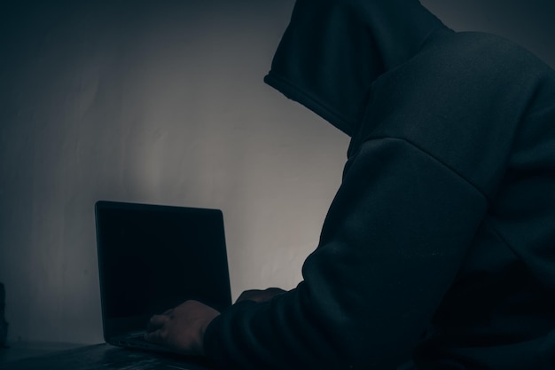 A hacker with a hood covering his head Sitting in a dark room using a laptop