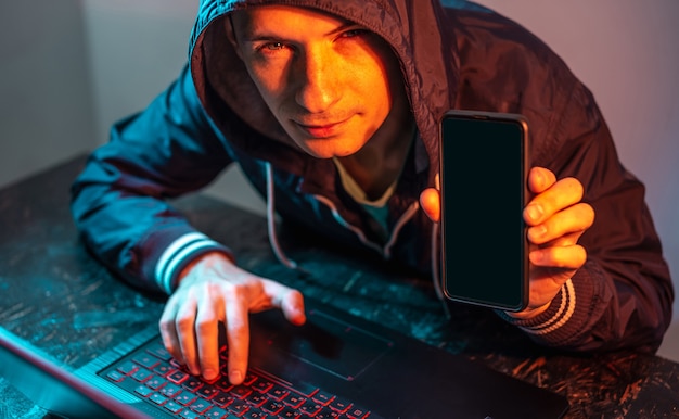 A hacker with a phone is typing on a laptop keyboard in a dark room under a neon light