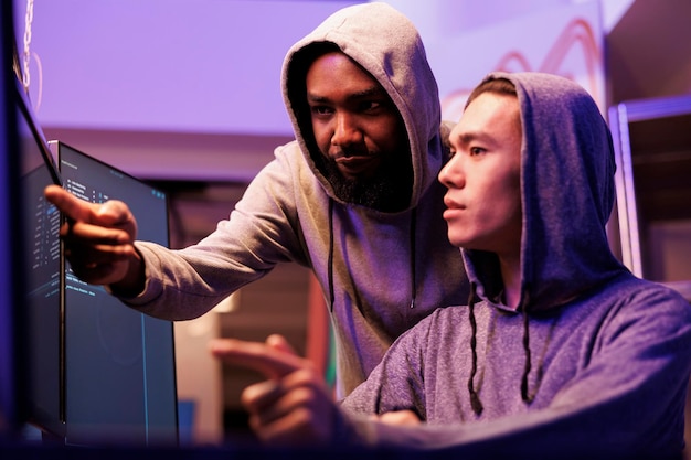 Hackers checking data after breaking database server together. African american criminal pointing at computer screen while breaking into network system with asian crime partner