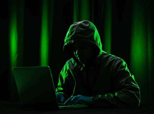 해커는 얼굴을 가리기 위해 후드를 착용합니다. 중요한 정보를 훔치는 해킹. 컴퓨터를 사용하여 맬웨어 바이러스 몸값을 배포하고 조직을 괴롭히십시오. 그는 네온 불빛이 있는 어두운 방에 앉아