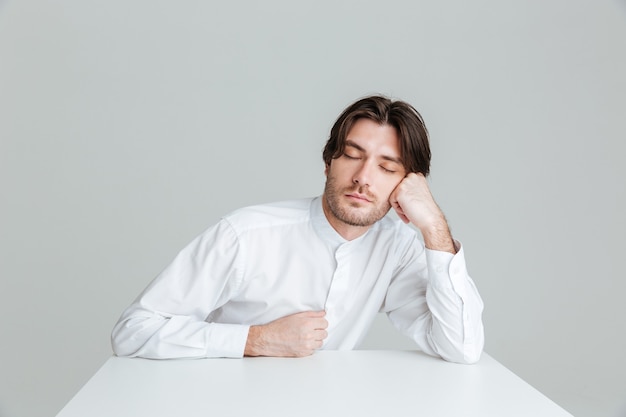 Hadsome young man in white shirt sleeping while sittng at the desk isolated on the gray wall