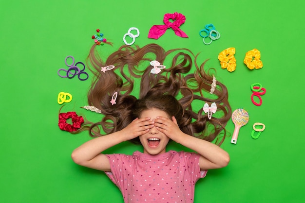 Hair accessories A happy little girl lies surrounded by elastic bands and hair clips and covers her eyes with her hands smiling broadly Hairstyles for children Green isolated background