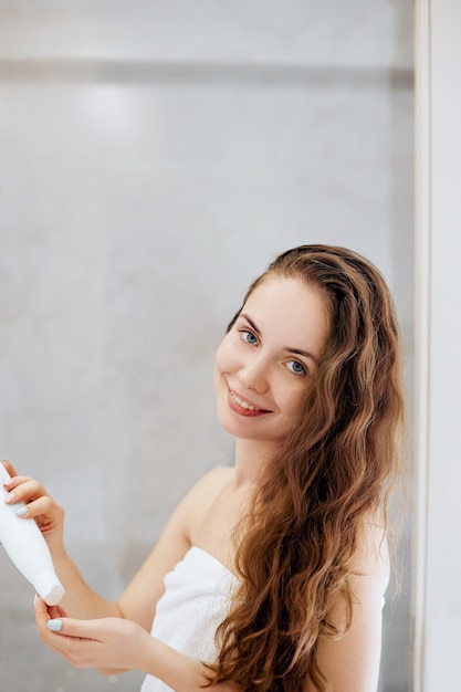 Hair. Beautiful young woman applying lotion for hair and smiling while standing in front of the mirror  in bathroom. Care hair and skin.  Girl uses protection moisturizing cream.