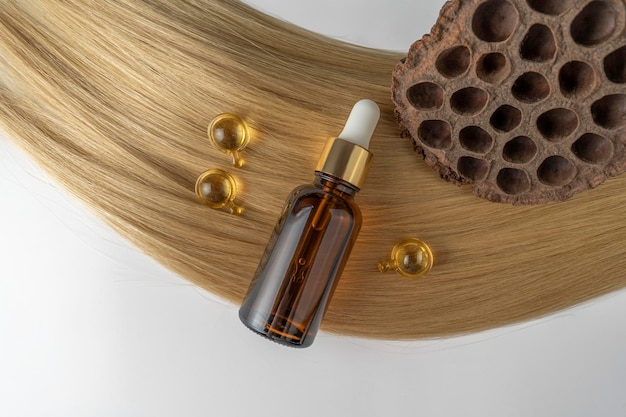 A hair care oil or serum in a brown dropper bottle and golden capsules lying on a blond hair strand product marketing mockup