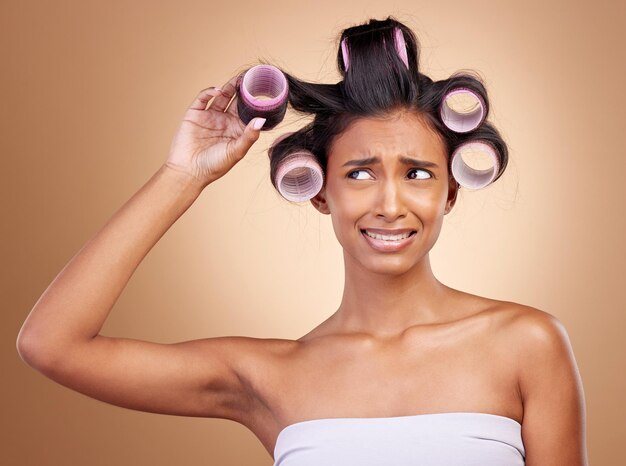 Hair care problem and a woman with rollers on a studio background for fail of curls Sad beauty and a young Indian model or girl with gear for a curly hairstyle mistake isolated on a backdrop