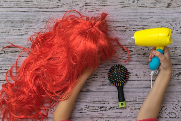 The hair dryer in the child's hand is aimed at a bright orange wig. Accessories for creating style and hair care.