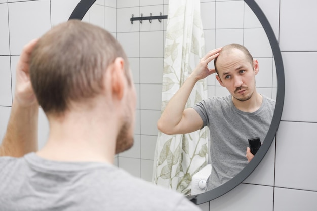 写真 抜け毛の問題ヘアクリッパーを持ってバスルームの鏡を批判的に見ている男性