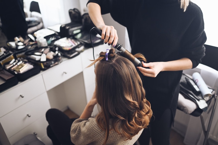 Photo hair stylist prepares beautiful young woman for event, makes curls hairstyle with a curling iron for client in beauty salon
