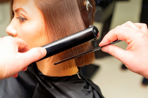 Hair stylist's hands straightening short hair of young brunette woman with flat iron and comb in a beauty salon