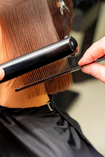 Hair stylists hands straightening short hair of young brunette woman with flat iron and comb in a beauty salon