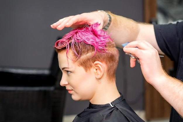 Стрижка окрашенных коротких розовых влажных волос молодой кавказской женщины парикмахером-мужчиной в парикмахерской