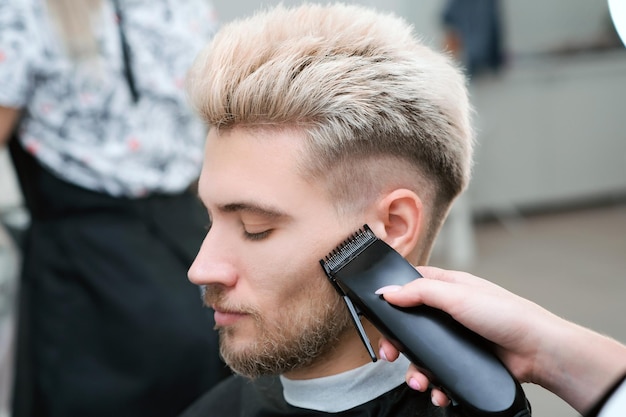 Haircut process of blond young man with hair trimmer in armchair in barbershop salon, barbershop concept for men and boys