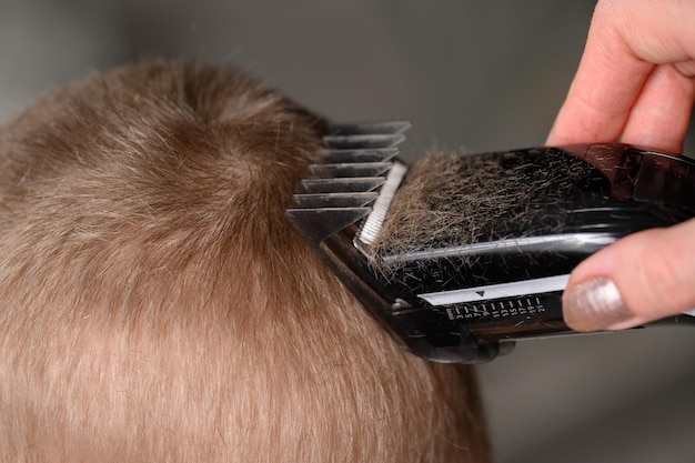 Фото Стрижки светлых волос для мальчика машинкой для стрижки крупным планом концепция ухода за волосамимать стрижет сыну волосы машинкой дома