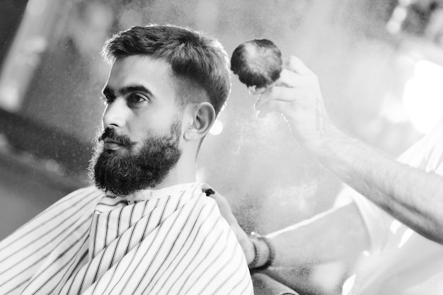 Hairdresser or barber does a hairstyle to a young man with a beard and mustache and pours talc on his hair