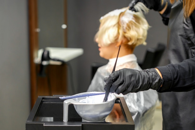 黒い手袋の美容師は美容院で白い色で若い女性の髪を染める