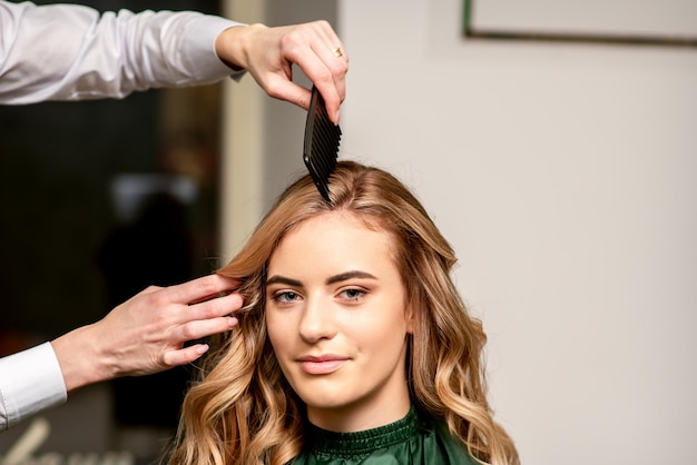 Parrucchiere che pettina i capelli lunghi della giovane donna caucasica che esamina la macchina fotografica e che sorride nel salone di bellezza