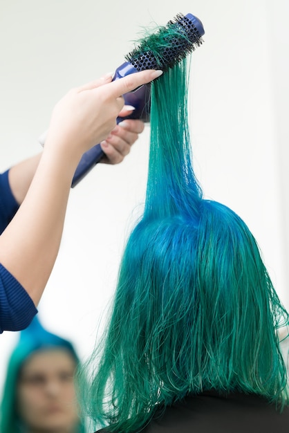 hairdresser combs and dries hair of emerald color with hairdryer after dyeing hair roots