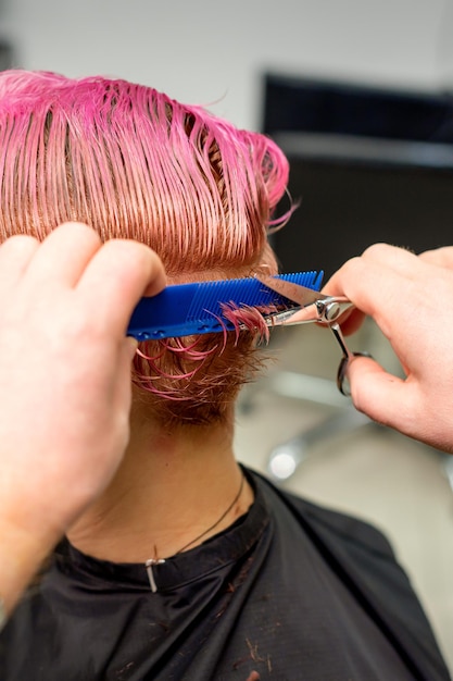 Парикмахер отрезает окрашенные влажные розовые короткие волосы молодой кавказской женщины, расчесывающей расческой в парикмахерской.