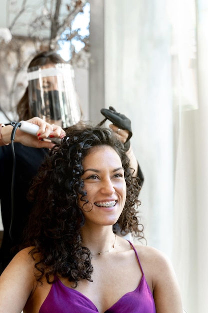 Parrucchiere in protezione facciale che arriccia i capelli di una cliente di bellezza in un salone