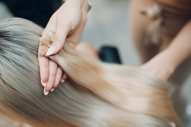 Hairdresser female making hair extensions to young woman with blonde hair in beauty salon. Professional hair extension strand of hair.