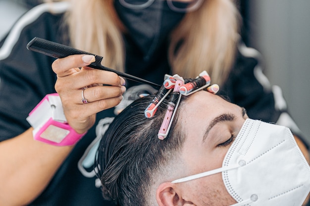 Parrucchiere che fissa i riccioli dei capelli di un giovane uomo caucasico con maschera usando il perno di un pettine