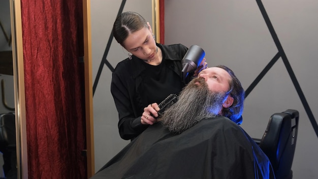 A hairdresser girl using a hair dryer to dry a man's long beard in a barbershop