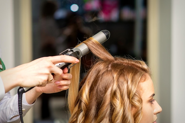 Il parrucchiere fa i ricci con un ferro arricciacapelli per la giovane donna con lunghi capelli castani in un salone di bellezza.