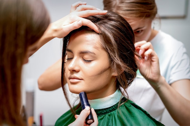 Il parrucchiere e il truccatore stanno facendo acconciatura e trucco per una bellissima giovane donna caucasica in un salone di bellezza