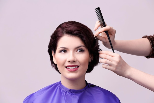 Hairdresser making beautiful haircut to young woman on color background