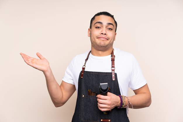 Hairdresser man in an apron isolated on beige wall having doubts with confuse face expression