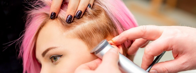 사진 미용사는 미용실에서 전기 면도기로 분홍색 머리로 여성 사원을 면도합니다.