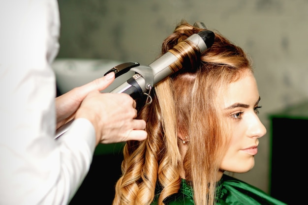 Hairdresser using curling tongs curls long brown hair on the young caucasian girl in a beauty salon