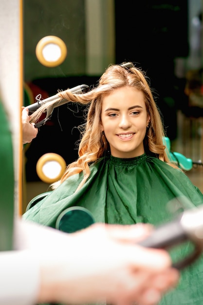 カーリングトングを使用している美容師は、美容院で若い白人の女の子の長い茶色の髪をカールします。
