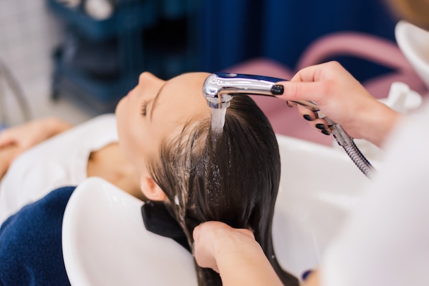 Hairdresser washing hair of woman female customer with a shower at the saloon applying shampoo conditioner on head hair