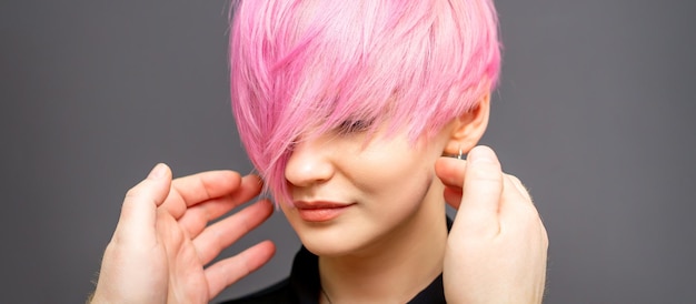 Hairdresser with hands is checking out and fixing the short pink hairstyle of the young white woman in a hair salon