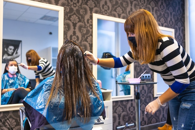 Hairdresser with mask applying the dye in front of the mirror to the client with mask. Reopening with security measures of Hairdressers in the Covid-19 pandemic, coronavirus