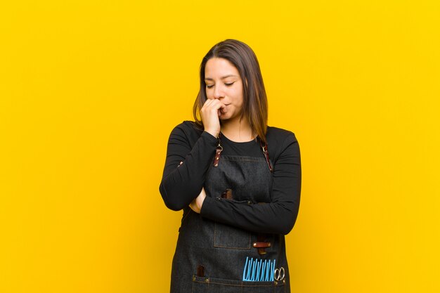 Hairdresser woman feeling serious, thoughtful and concerned, staring sideways with hand pressed against chin against orange wall