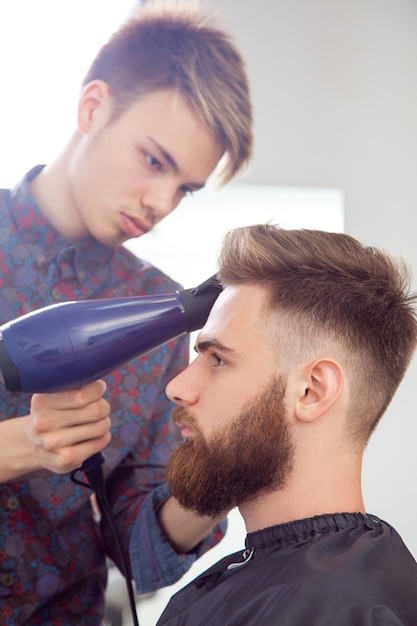 Hairstyling process. Close-up of a barber drying hair of a young bearded man