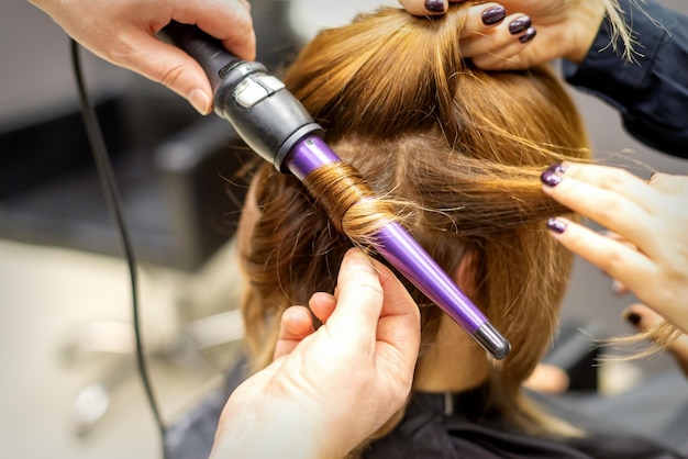 The hairstylist makes curls hairstyle of long brown hair with the curling iron in hairdresser salon close up