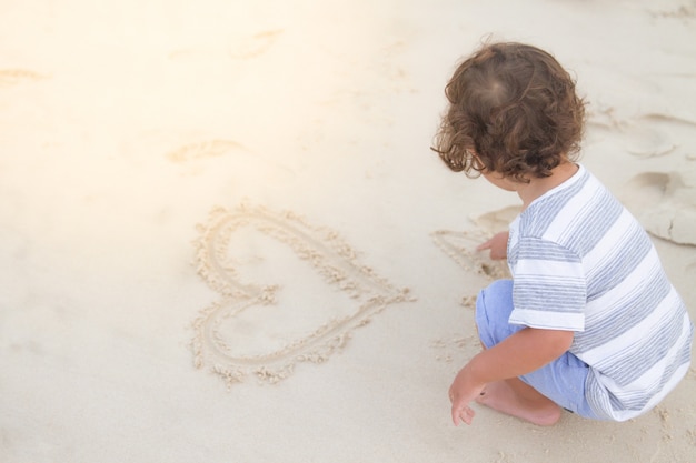 a hairy boy is drawing a heart on white sand beach