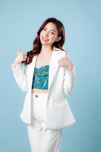 Half body of confident elegant beautiful Asian Business woman wearing white suit smiling and showing thumbs up on isolated blue background Young shopaholic female on copy space