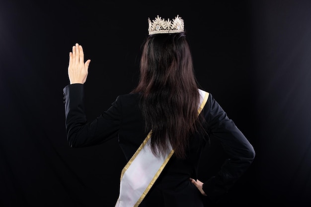 Half body portrait of Miss Pageant Beauty Contest in Asian smart suit blazer shirt dress with Silver Diamond Crown Sash fashion make up smile wave hand studio lighting black background isolated