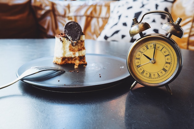 Half chocolate cake and alarm clock on black table.