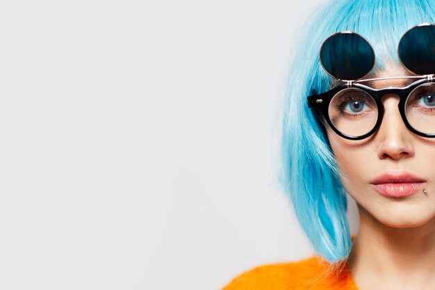 Half face concept; close-up studio portrait of young pretty young girl with blue hair, wearing round sunglasses and orange shirt on white background with copy space.