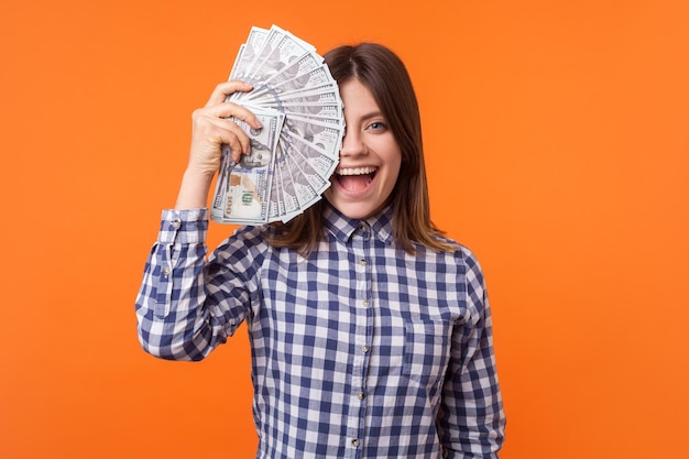 Half face portrait of amazed attractive brunette woman wearing checkered shirt holding dollars and looking at camera with open mouth lottery winner indoor studio shot isolated on orange background